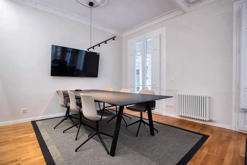 Sala de reuniones con mesa negra, sillas claras, pantalla y suelo de madera